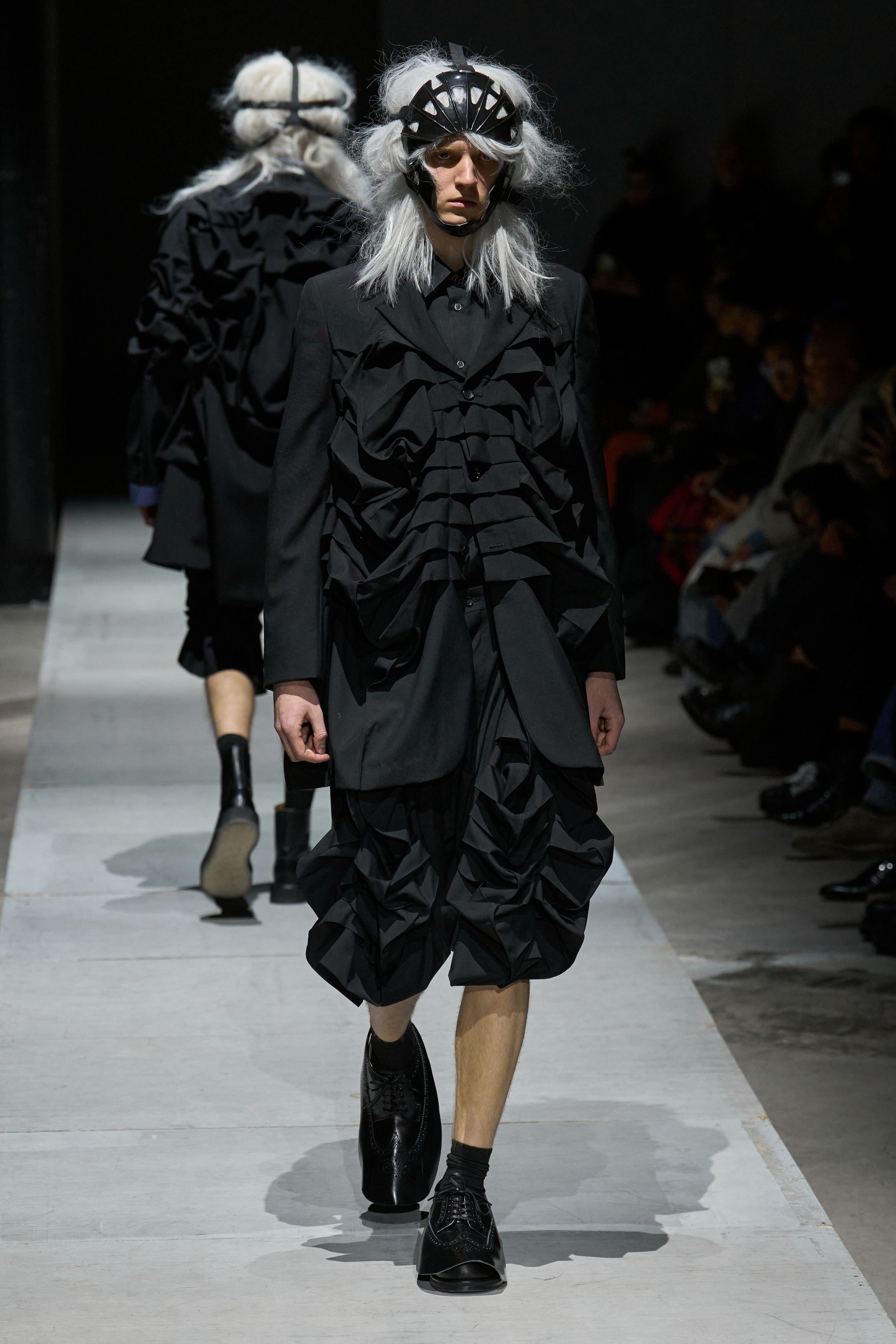 Comme des Garçons Homme Plus 2026秋冬以极端造型继续挑战“男性身体”的社会想象。面具、假发、解构西装与异化比例营造出近乎仪式化的压迫氛围，黑白灰成为思想载体而非装饰。这是一场关于权力、恐惧与集体意识的时装实验，服装不再服务于穿着，而是成为观念的武器-顽石频道趋势研究